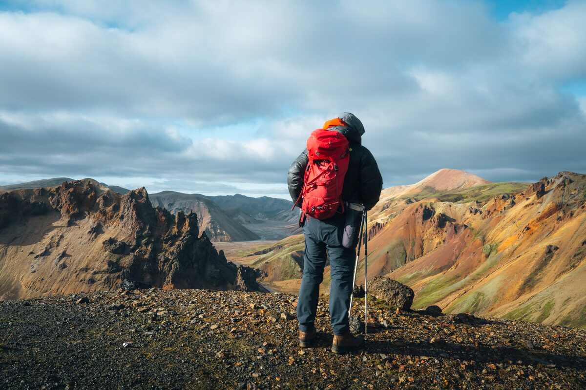 Hiker with a red backpack overlooking Iceland’s colorful mountain landscape under a cloudy sky.