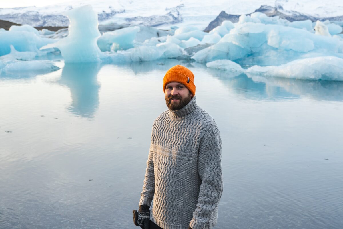 Man wearing a gray Icelandic sweater and orange beanie standing by floating icebergs at a glacier lagoon.