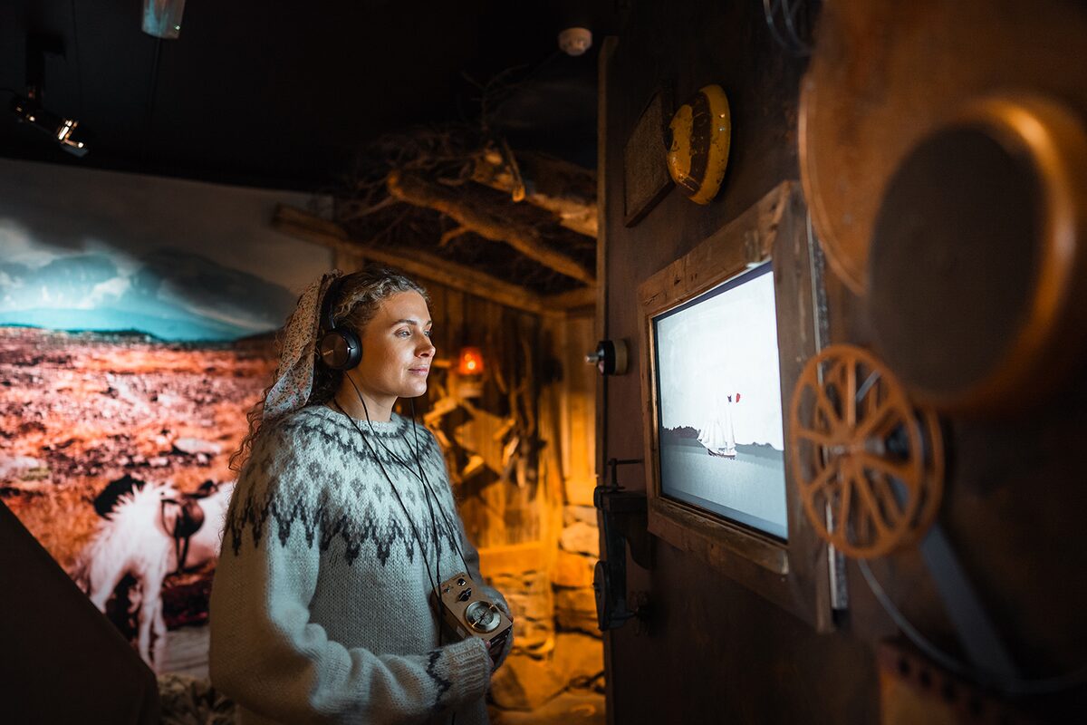Person wearing a patterned Icelandic sweater and headphones inside a museum-style exhibit, looking at a screen.