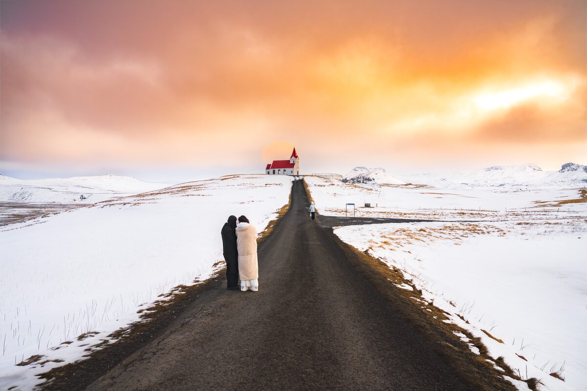 Couple standing on road by Ingjaldsholskirkja church during bright sunset in winter.