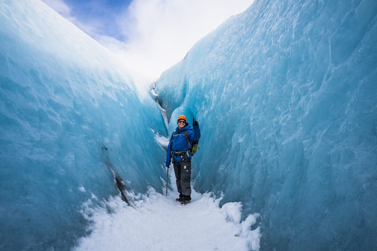 Happy man in orange helmet standing between walls of blue ice holding up #1 hand