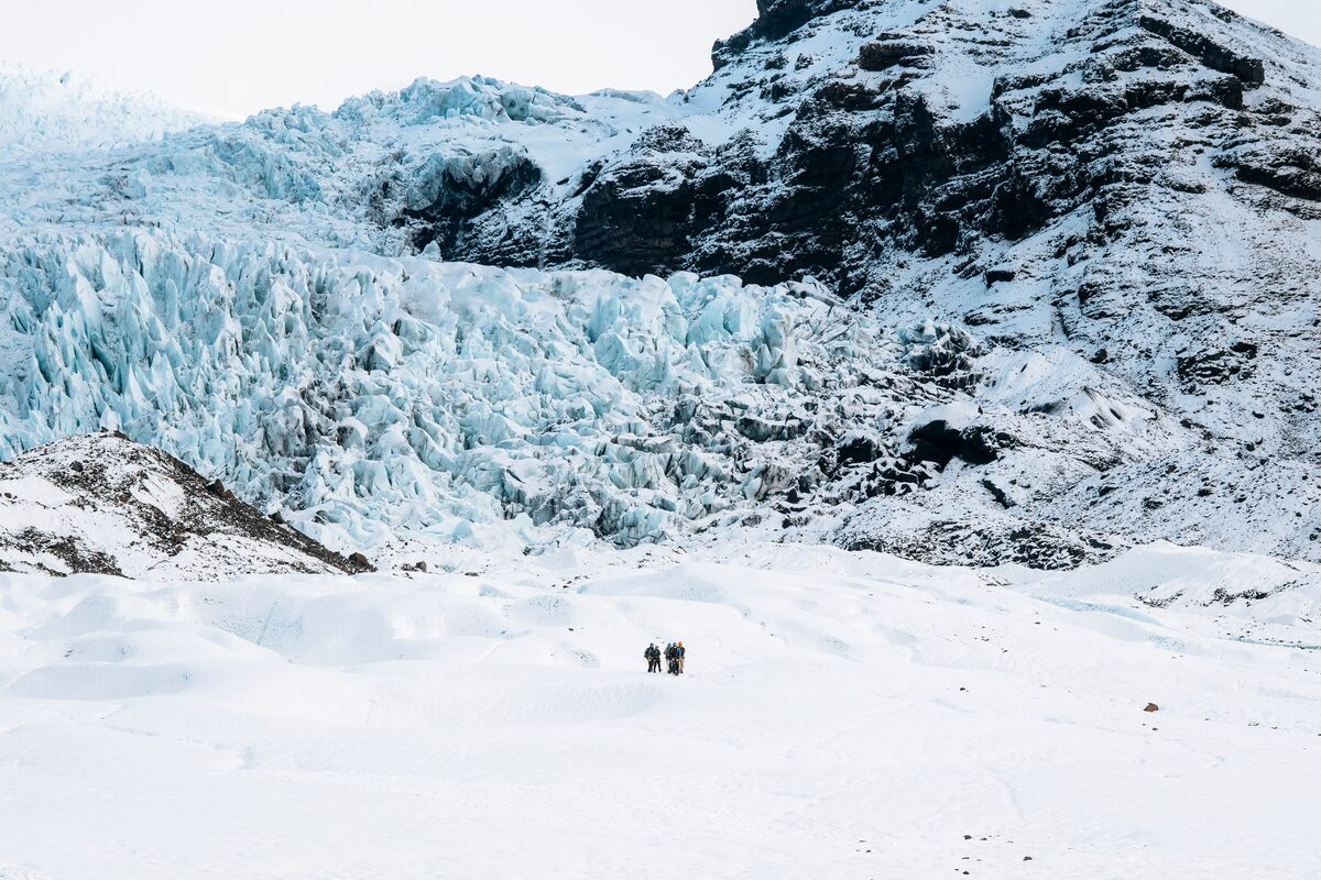 Distance shot of group looking small in front of jagged blue ice and black & white mountain.