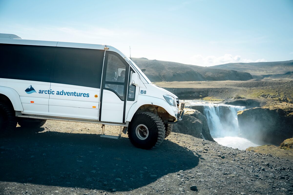 Arctic Adventures bus parked near a waterfall in Iceland, with a rocky landscape and mist in the background.