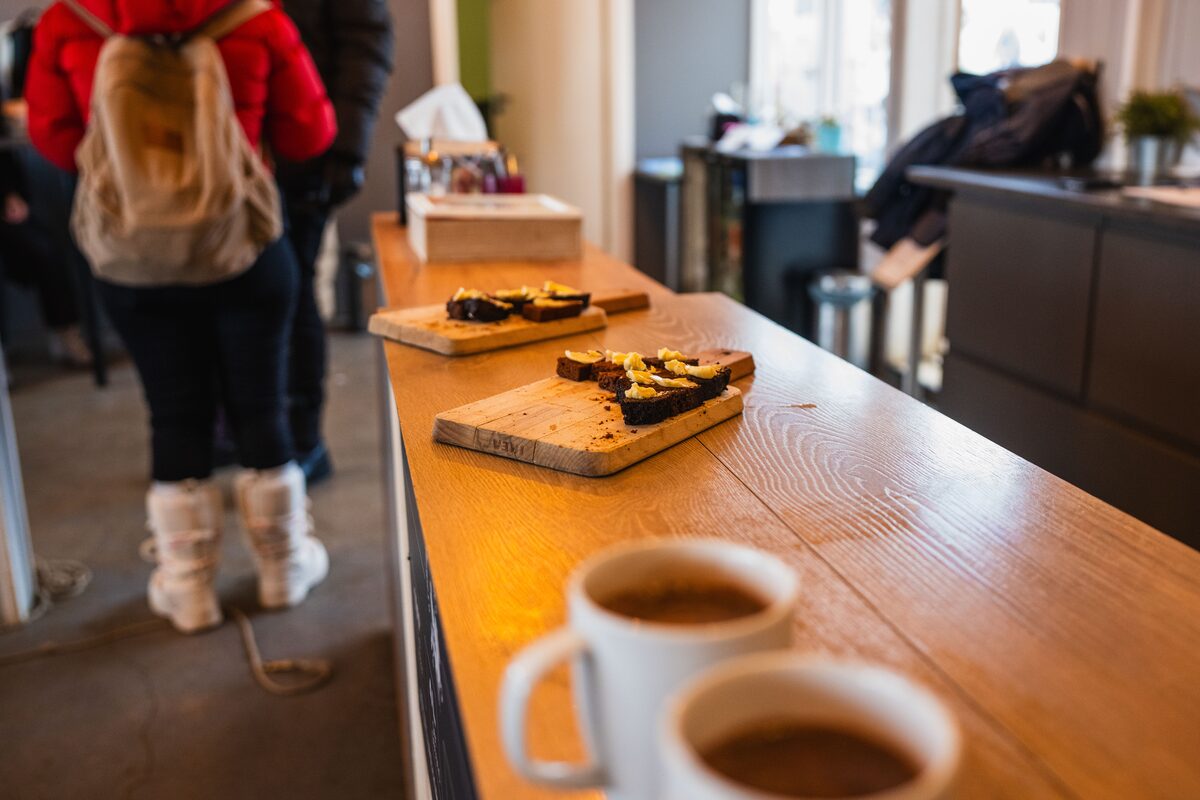 Coffee cups on a cafe counter, with snacks on boards and customers in the background.