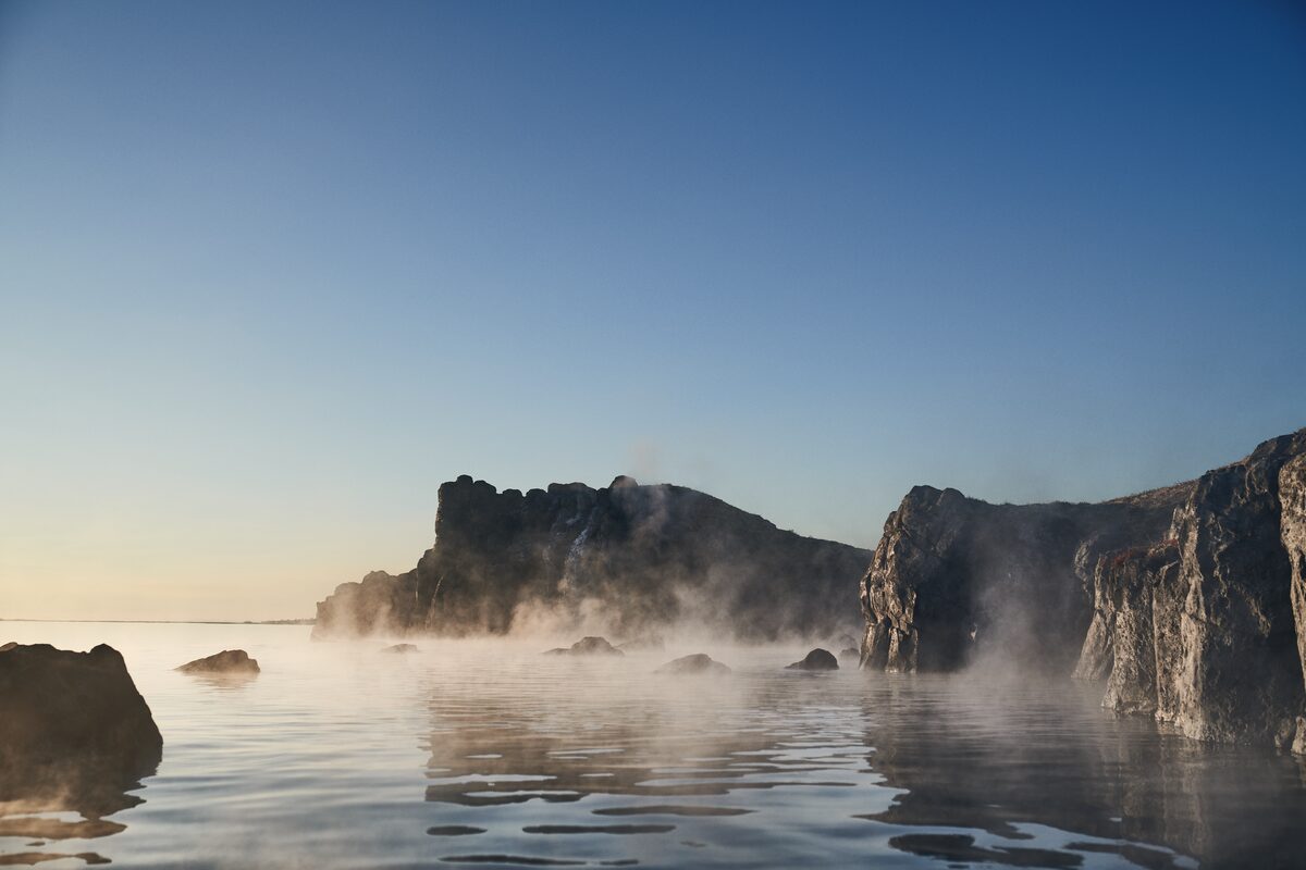 Steam rising over Sky Lagoon's geothermal waters with dark lava cliffs at dusk, Kópavogur, Iceland.