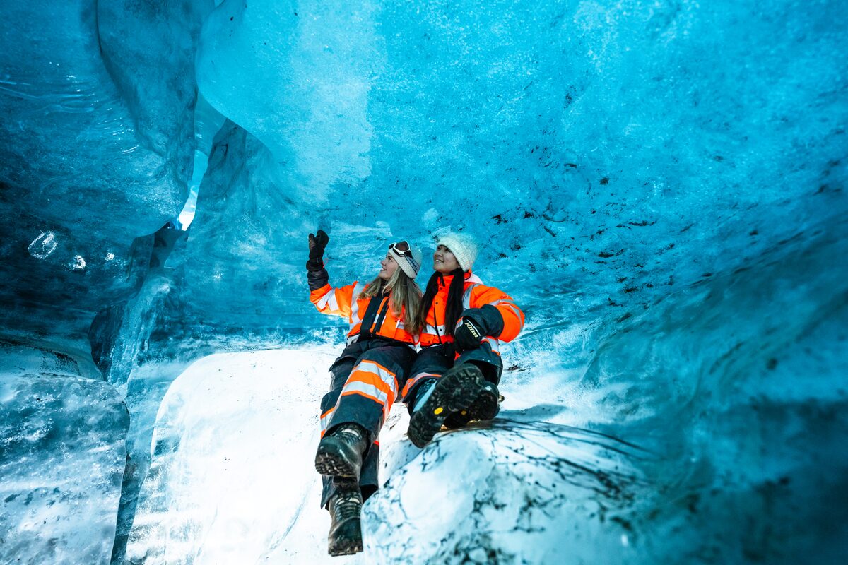 Two people in orange suits inside a blue ice cave in Iceland.