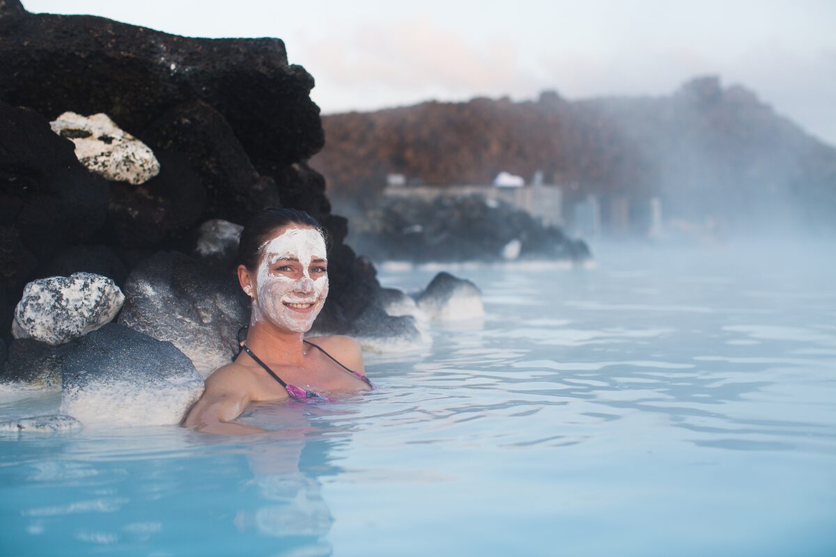 Woman smiling in the Blue Lagoon with a white silica mud mask on her face, surrounded by black lava rocks.