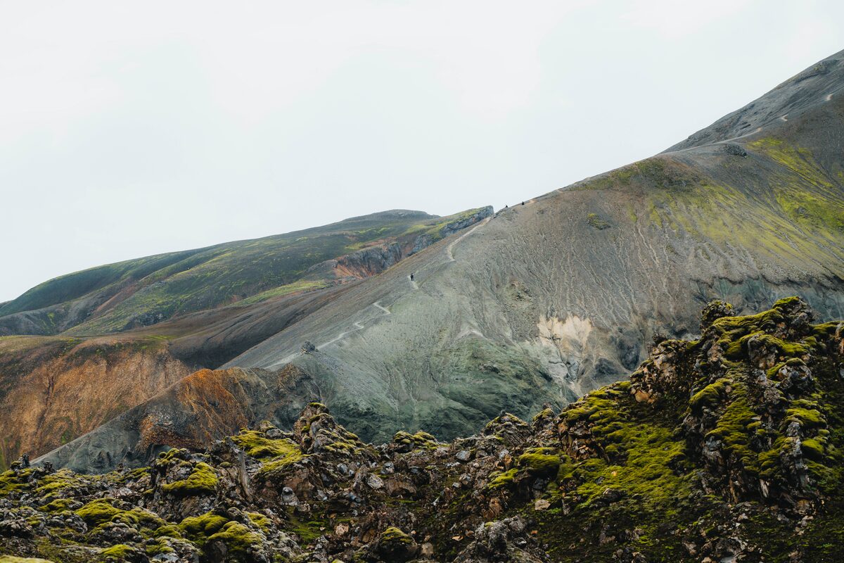 Hikers on a ridge trail in Landmannalaugar with Bláhnúkur's dark slopes and colorful rhyolite mountains behind.