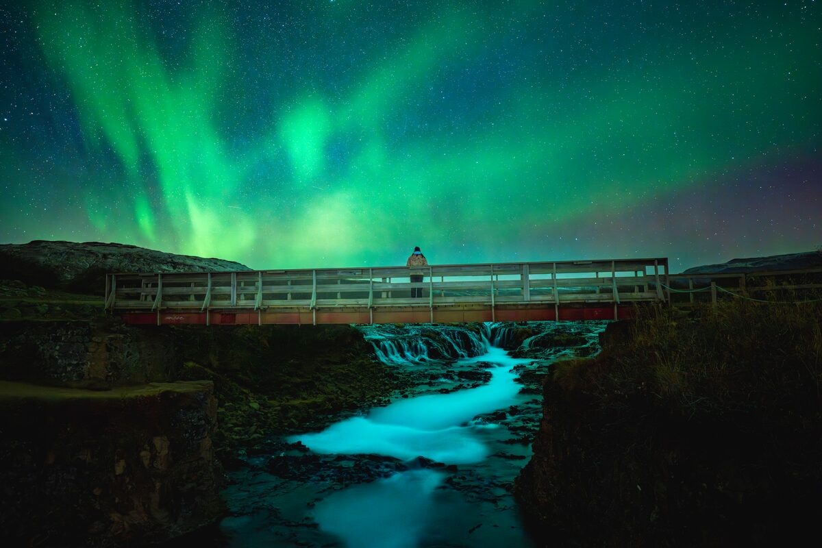 Person on a bridge above Brúarfoss waterfall under green Northern Lights in southwest Iceland.