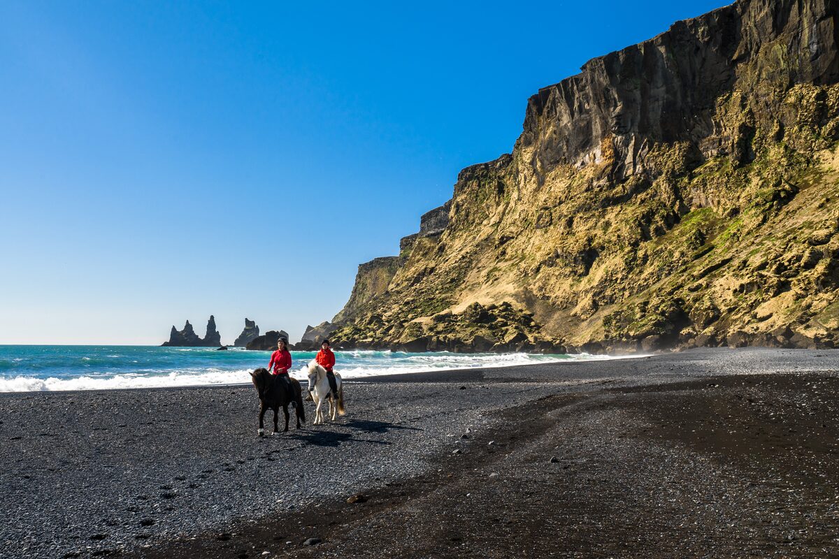 Two horseback riders on a black sand beach in South Iceland with sea stacks in the distance.