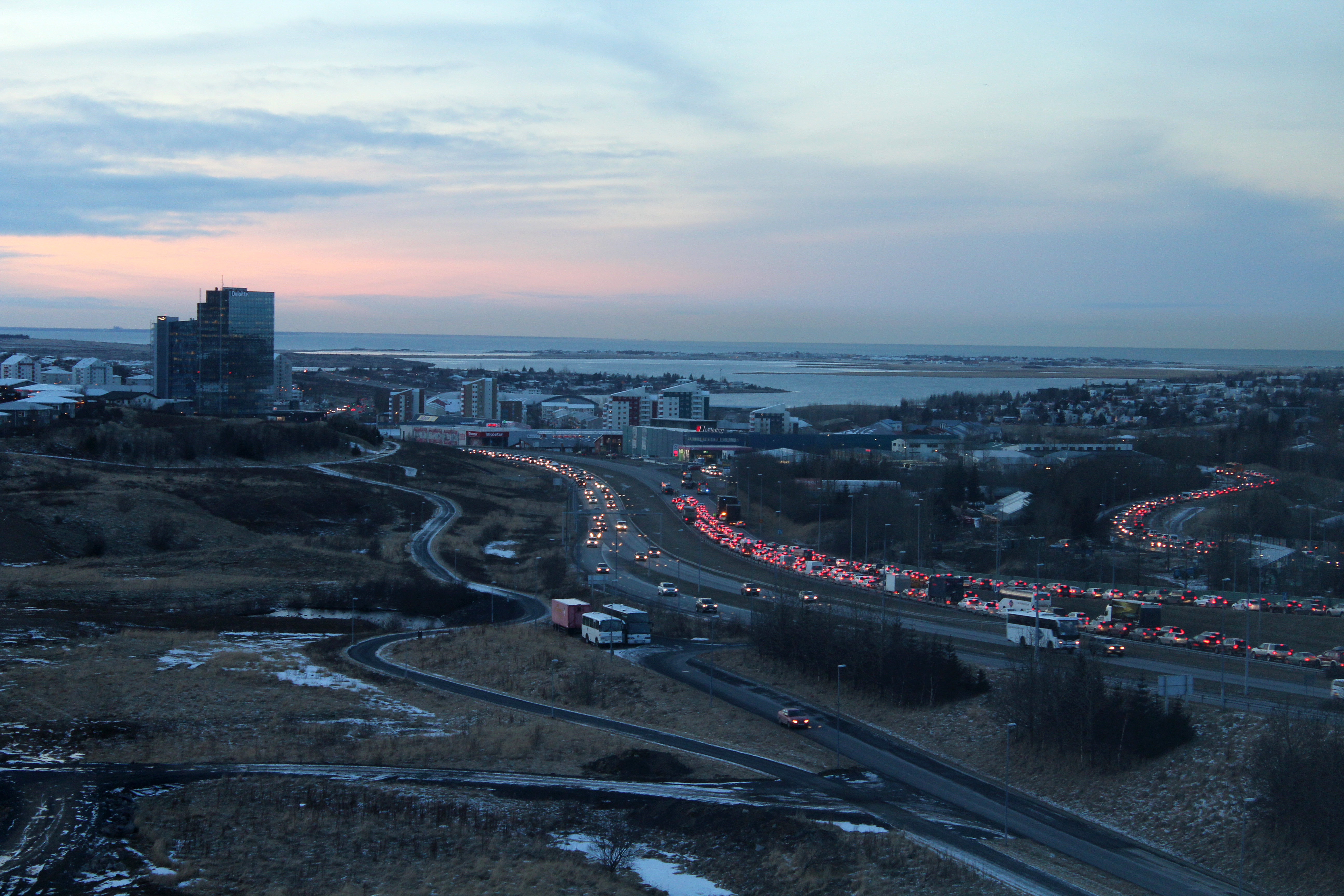 Reykjanesbraut traffic at dusk passing Smáratorg Tower in Kópavogur with Faxaflói Bay behind.