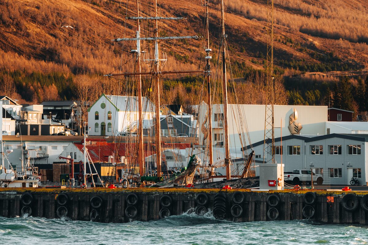 Tall-masted wooden boats at Húsavík harbor with Húsavíkurfjall hill behind in evening light.