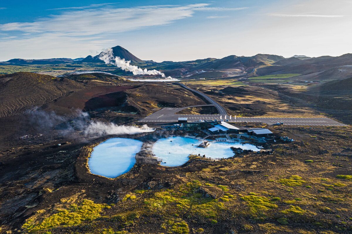 Mývatn Nature Baths with milky-blue geothermal pools, bathhouse, and steaming geothermal field and mountains behind.