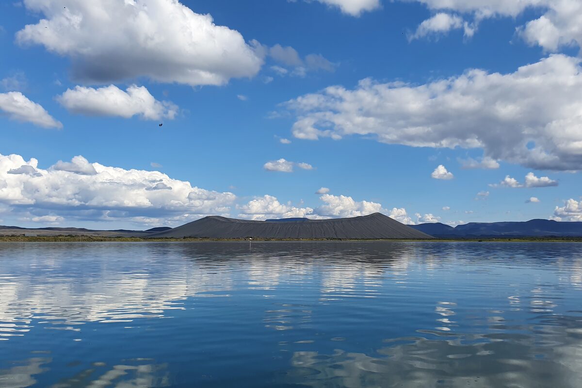 Flat-topped Hverfjall crater across calm Lake Mývatn under blue sky with white cumulus clouds.