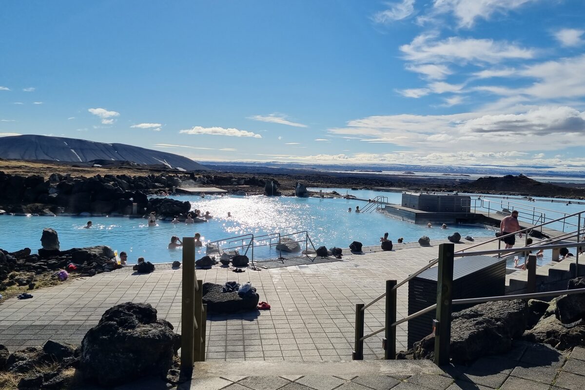 Mývatn Nature Baths with milky-blue geothermal pools, bathhouse, and steaming geothermal field and mountains behind.