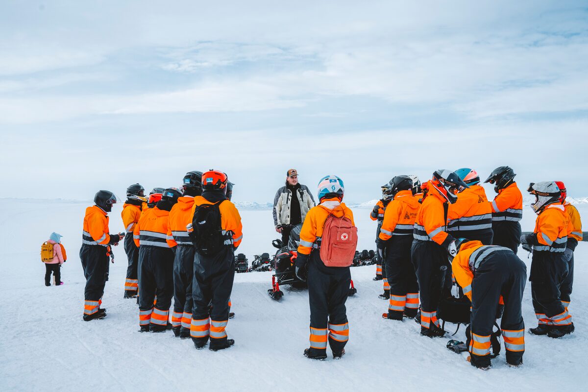 A guide briefing a group of riders in orange suits standing around snowmobiles on a glacier.