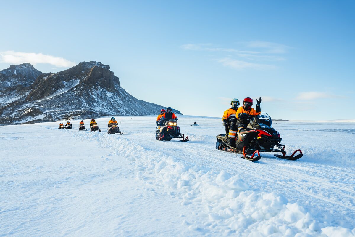 A line of snowmobiles riding across Vatnajökull glacier with ash-streaked snow and mountains. 