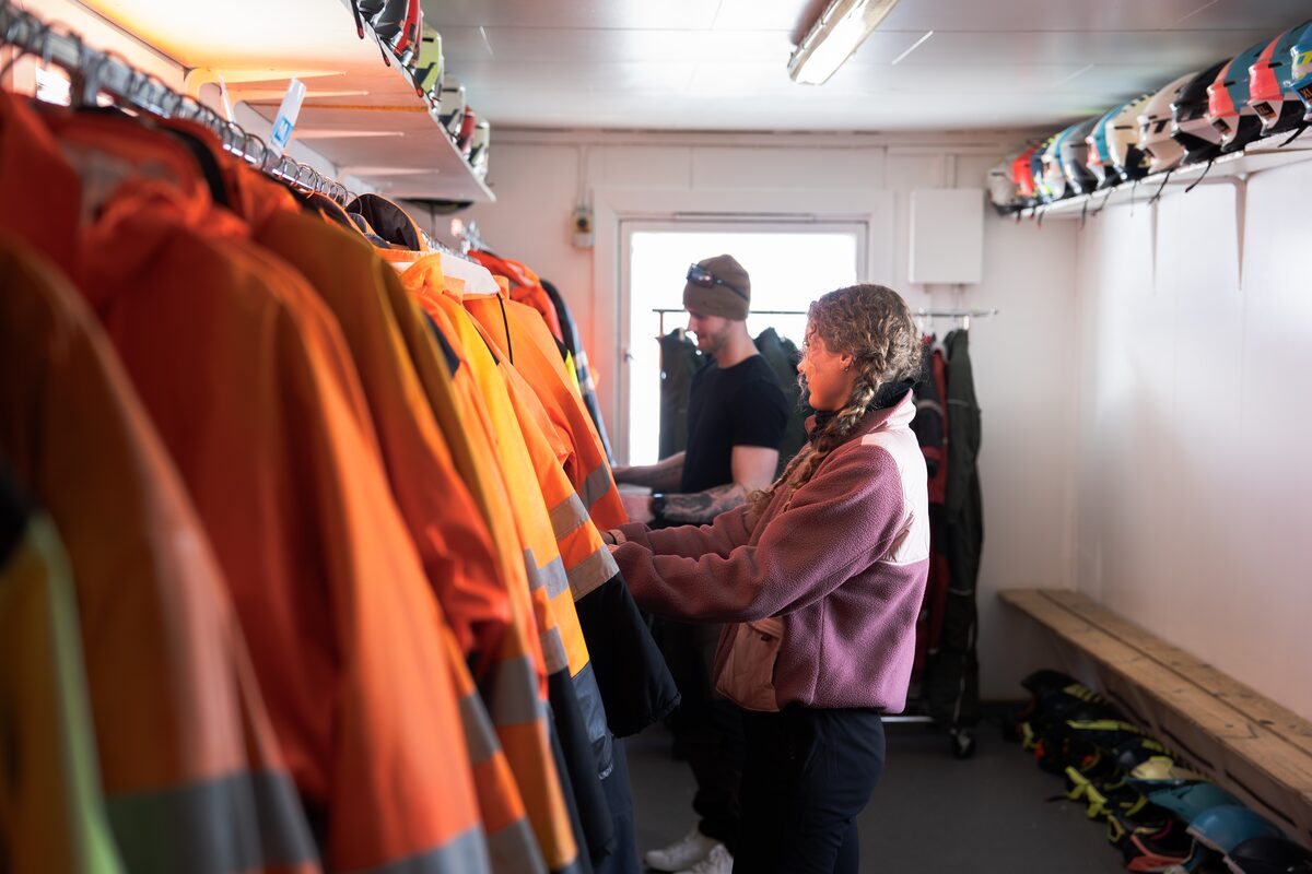 Two travelers picking out insulated suits and gloves from racks inside a gear fitting room.  