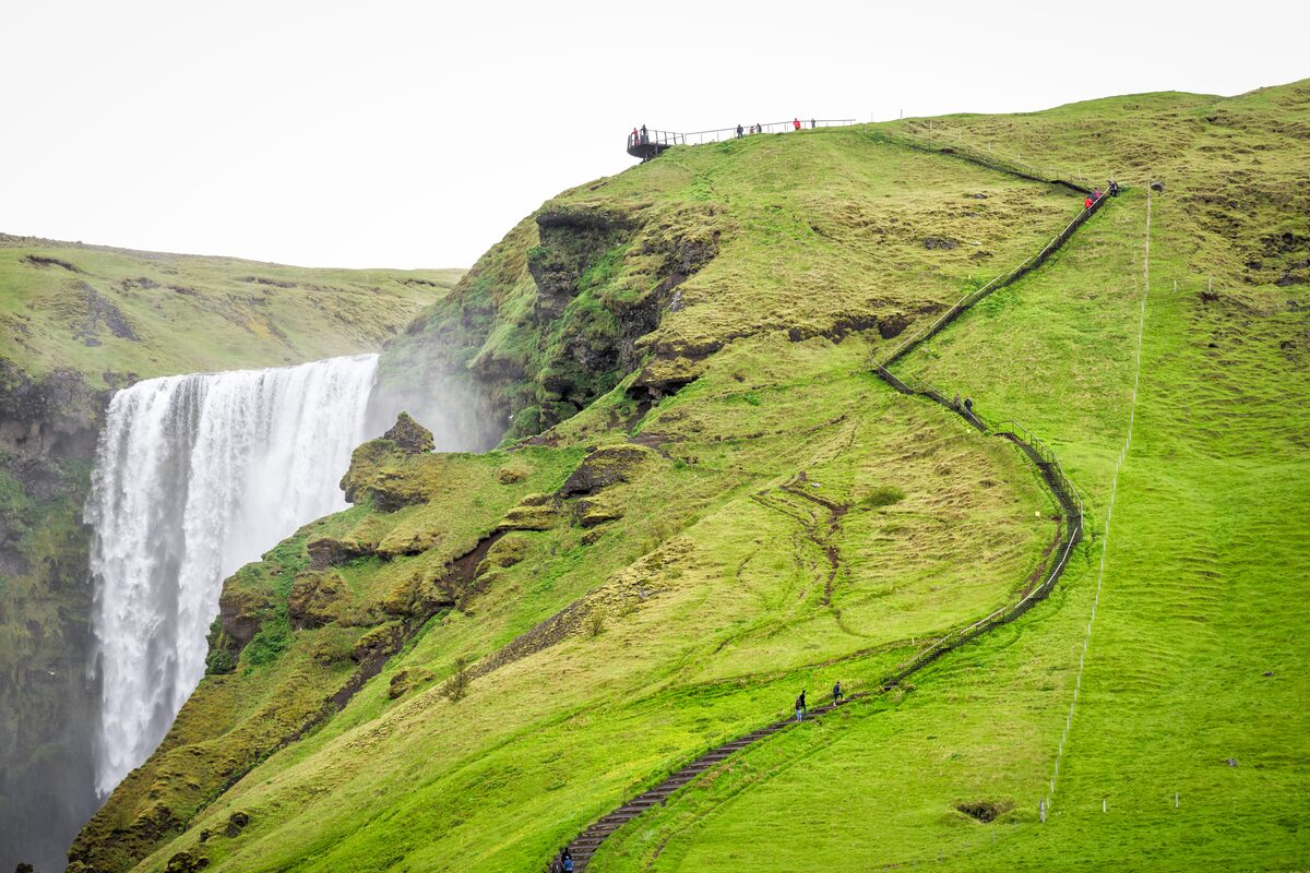 Staircase with handrail climbing the green cliff beside Skógafoss waterfall to a viewing platform. 