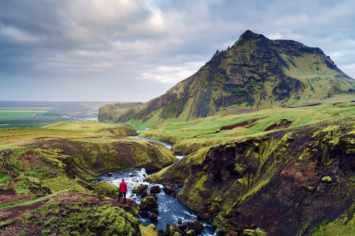 Hiker in red jacket above a small cascade in the Skógá river canyon, with green peaks beyond.