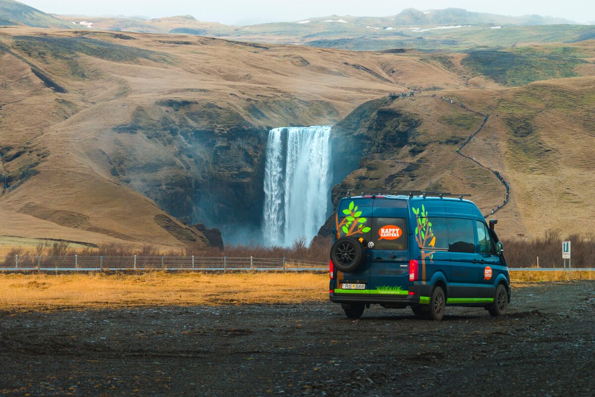 Campervan parked on gravel at Skógafoss waterfall, with cliffside staircase visible on the right.