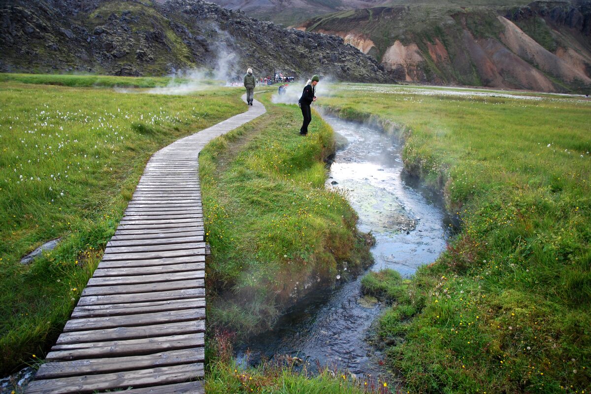 Visitors walking along a wooden boardwalk beside a steaming stream at Landmannalaugar, Iceland.