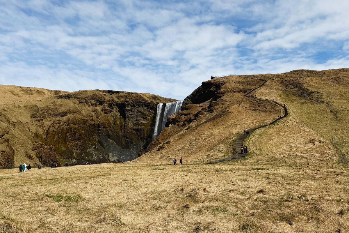 Staircase with handrail climbing the green cliff beside Skógafoss waterfall to a viewing platform. 