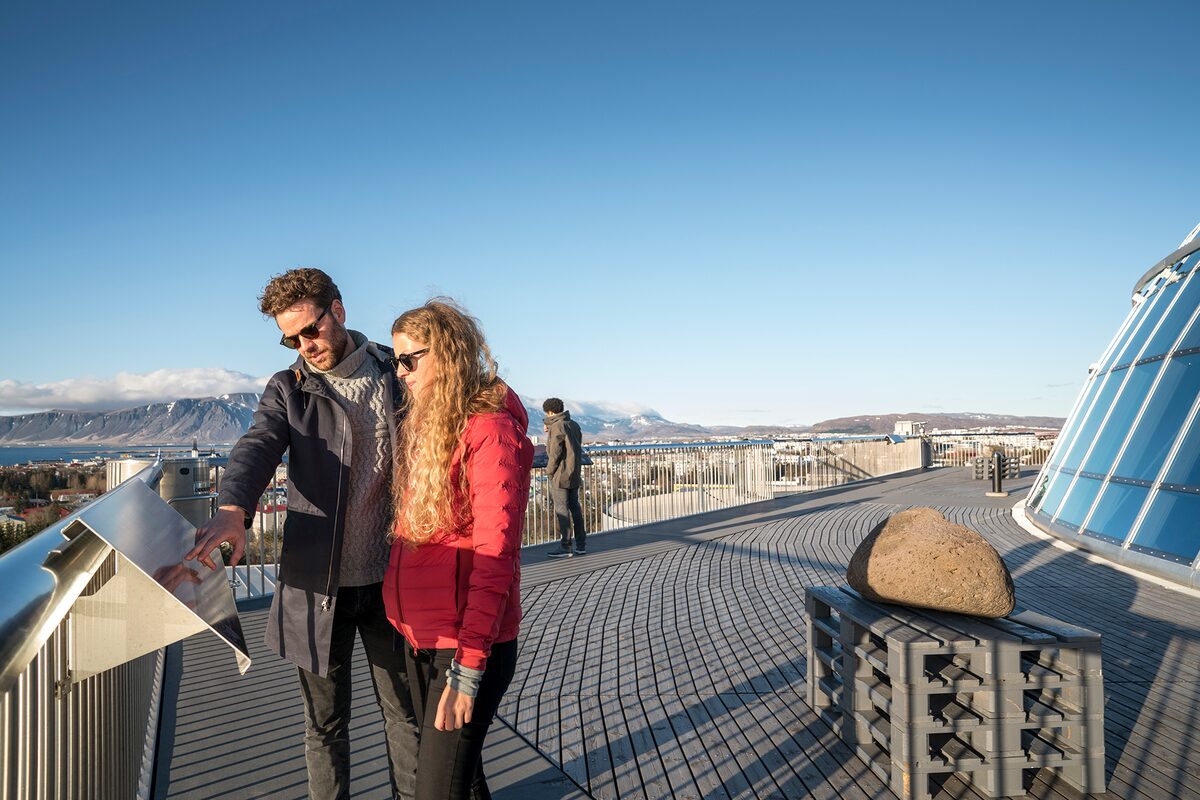 Couple at an interpretive panel on Perlan's observation deck with Mount Esja and Reykjavík behind.