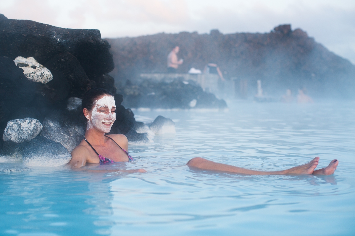 Person with a silica mud mask floating in the Blue Lagoon, lava landscape in the background.