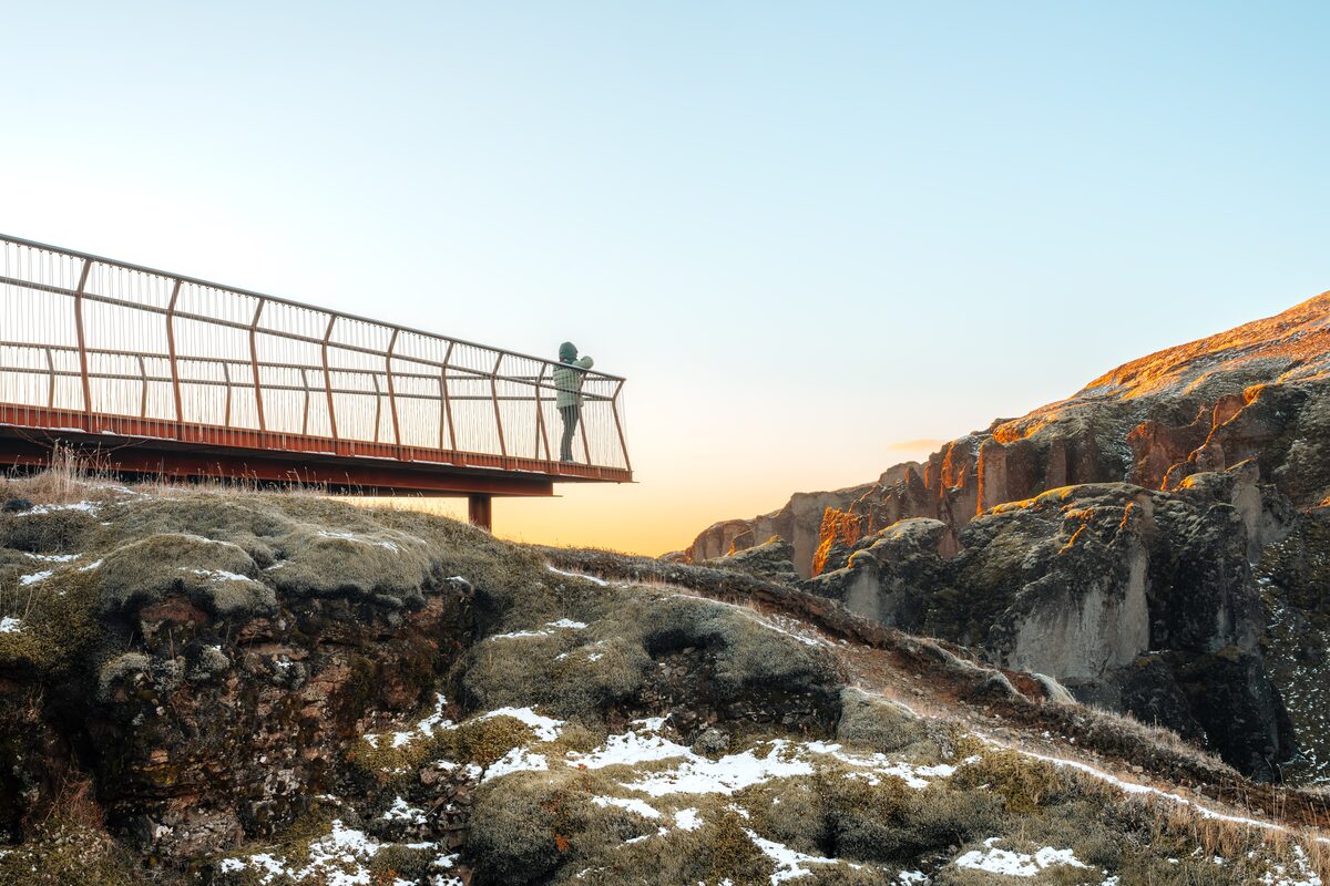 Person on a viewing platform over a mossy Fjaðrárgljúfur canyon with patchy snow at sunrise.