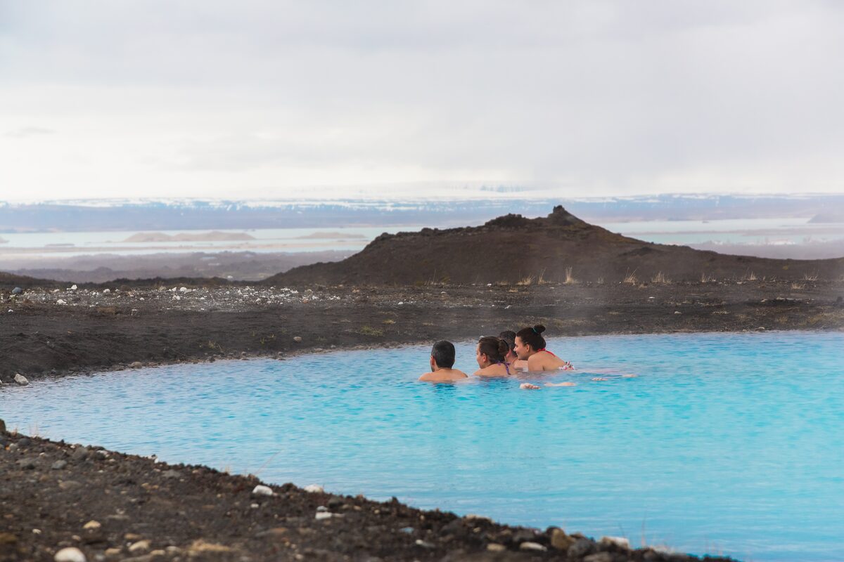Three bathers in milky-blue water with black lava and distant mountains beyond.