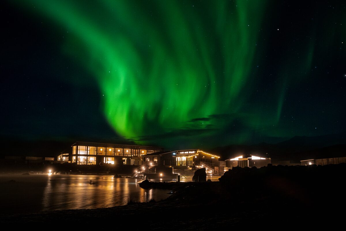 Green Northern Lights over the lit Mývatn Nature Baths building and lagoon at night.