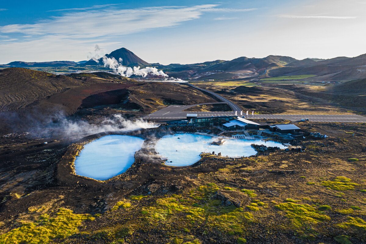 Aerial of Mývatn Nature Baths lagoon and spa building in a lava field, with a mountain behind.