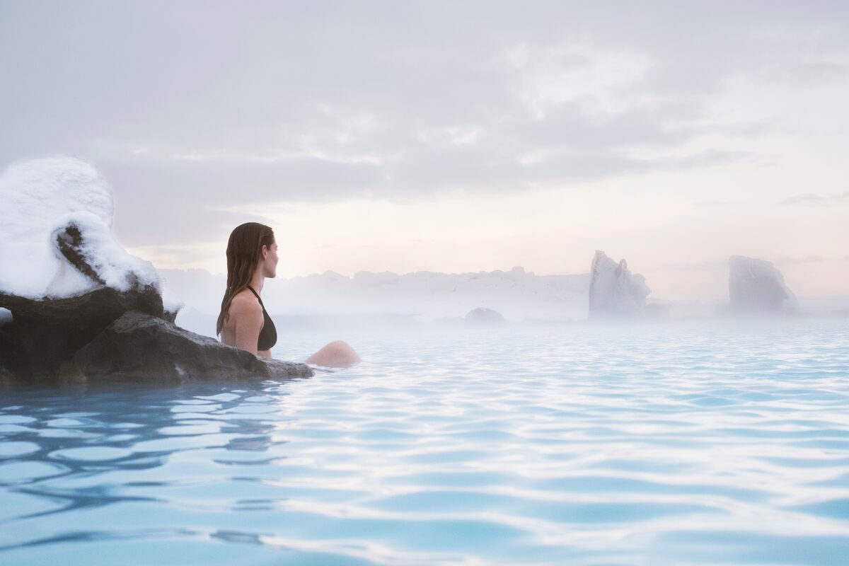 Woman sitting alone in milky-blue water with snow on black rock and mist over the surface.