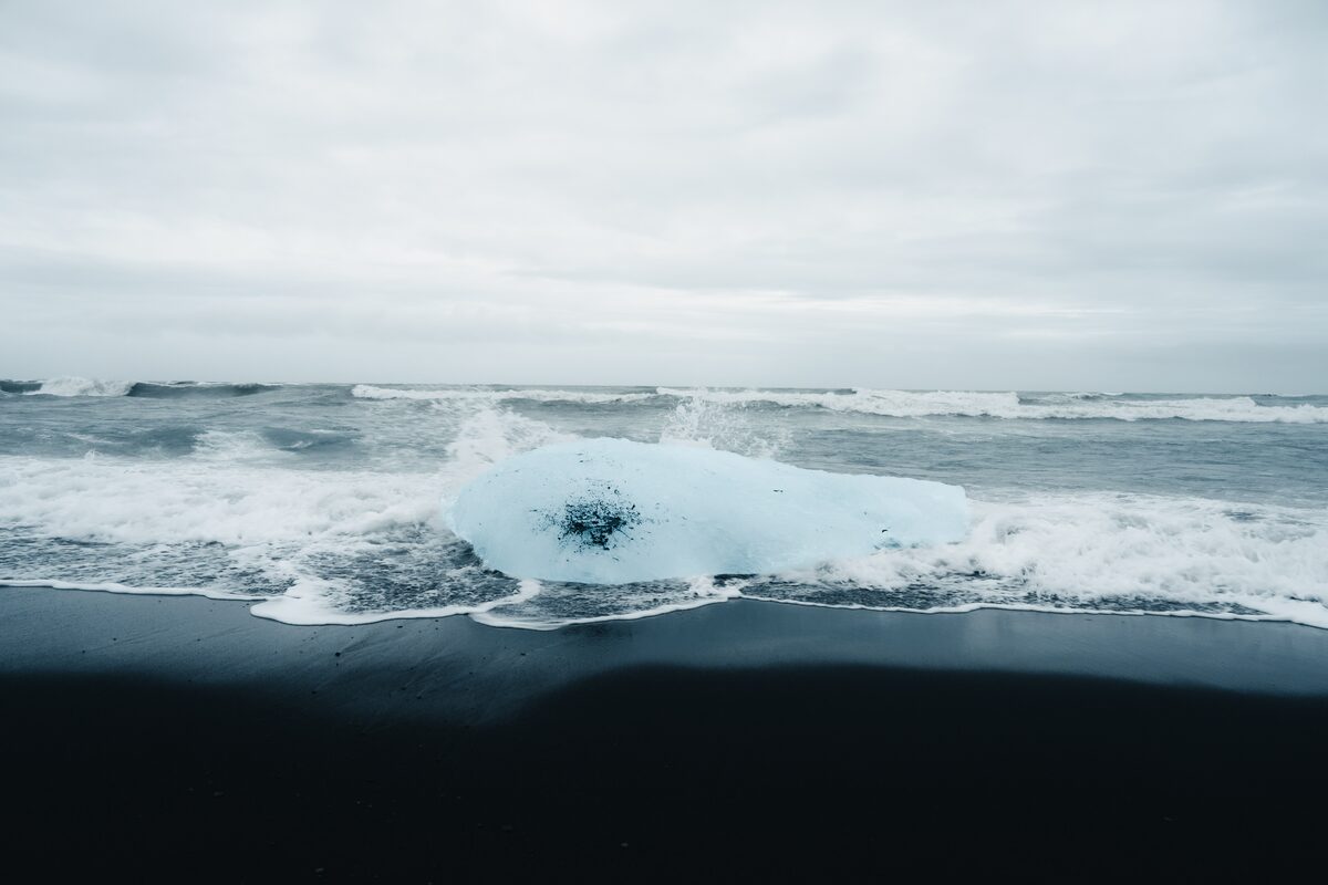 Single clear ice block against black sand and white sea foam.
