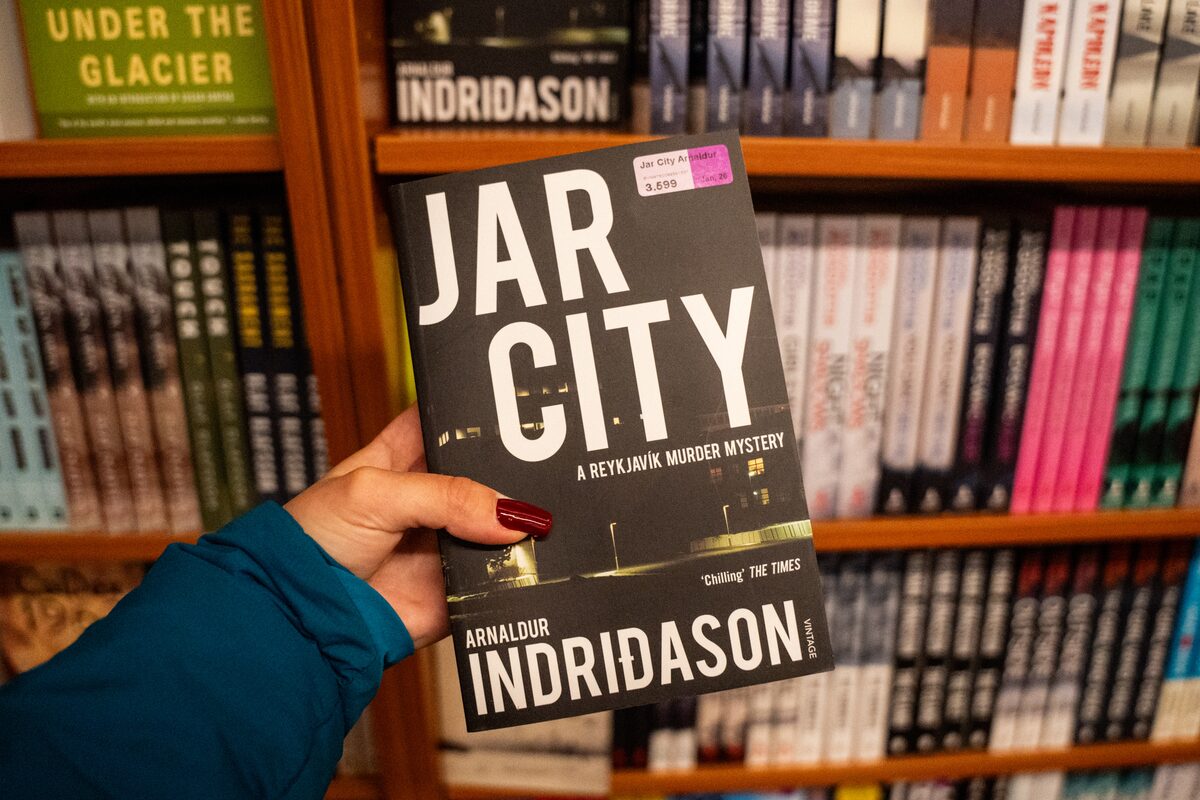 Person holds the crime novel Jar City in front of a shelf of Indriðason titles.