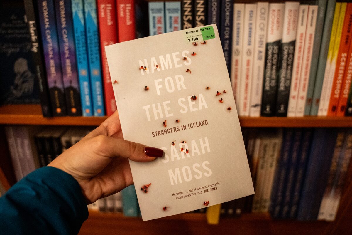 Person holds Sarah Moss's memoir Names for the Sea in front of a bookshelf.