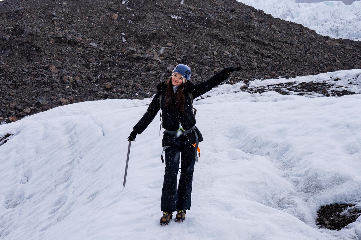 Woman in a helmet holding an ice axe on a snowy glacier in Iceland, one arm raised.