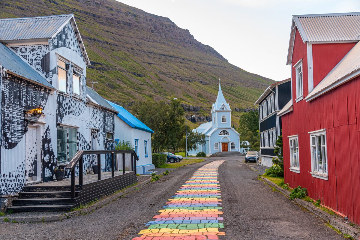 Rainbow-painted street leading to a blue church in Seyðisfjörður, with red and white houses alongside. 