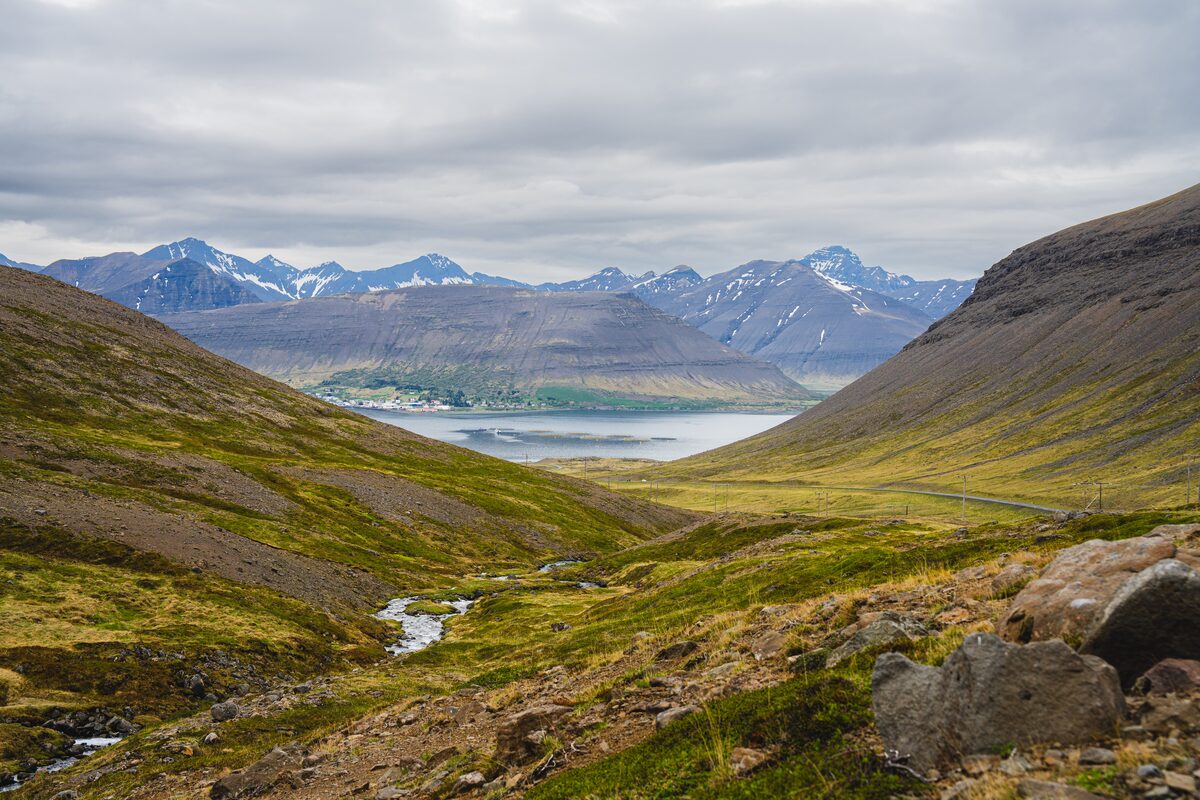 Green glacial valley and stream opening onto a fjord backed by snow-streaked mountains in the Westfjords.