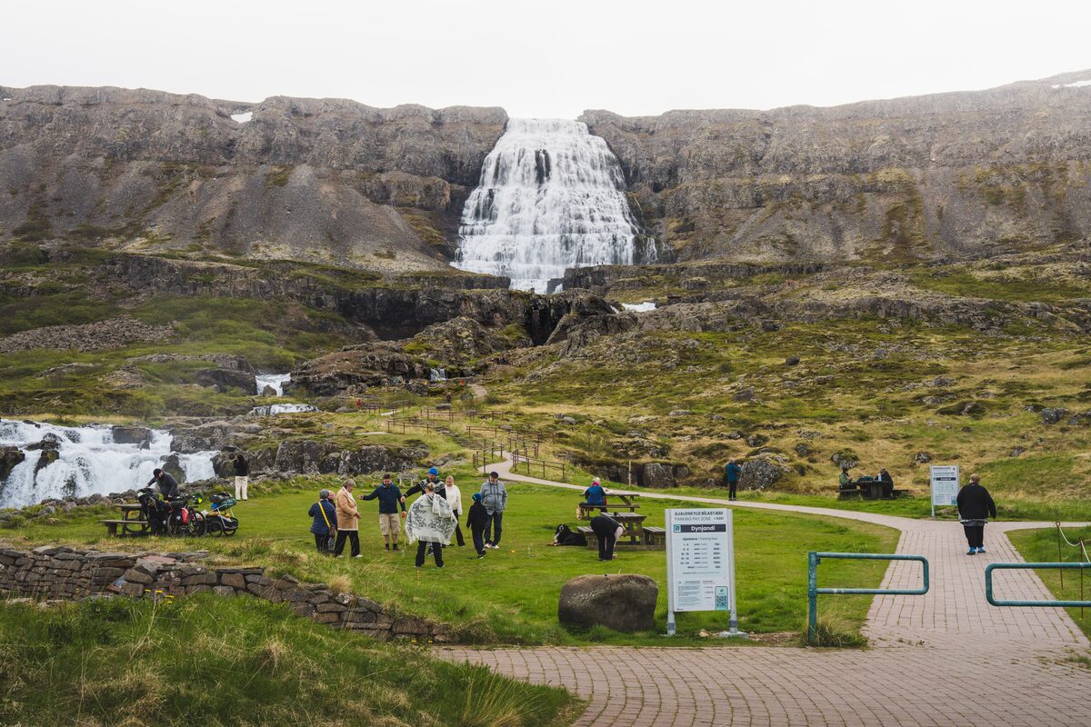 Dynjandi waterfall drops over a rocky cliff above paths, signs, and visitors in the Westfjords.
