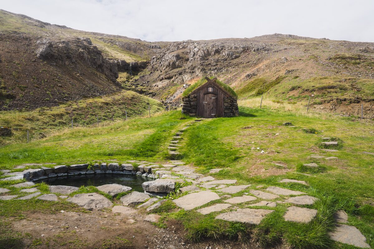 Turf-roofed hut beside a stone-lined natural hot pool on a grassy slope in Iceland