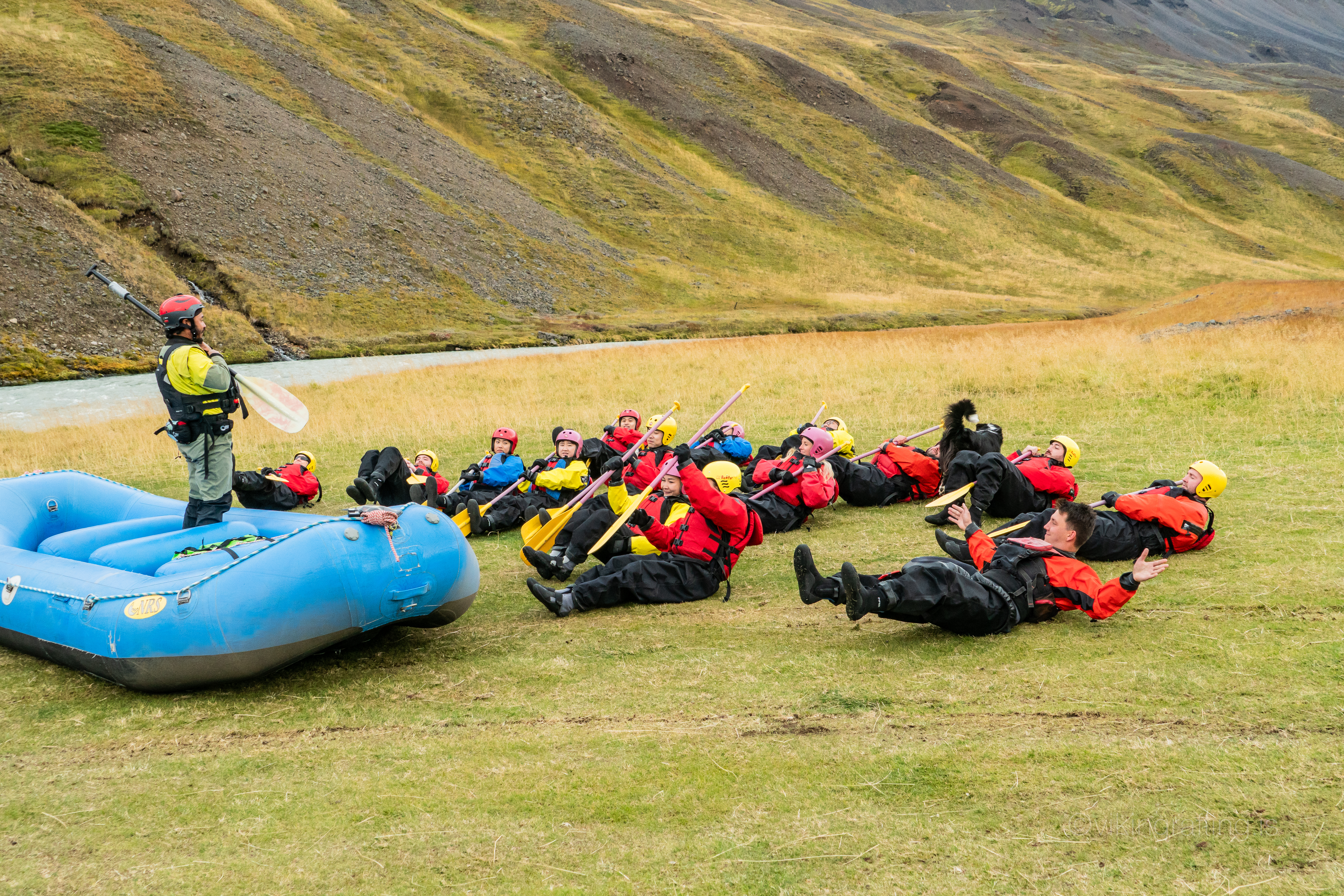 Rafters in helmets and suits lying on the grass holding paddles, with a guide and raft on the riverbank.