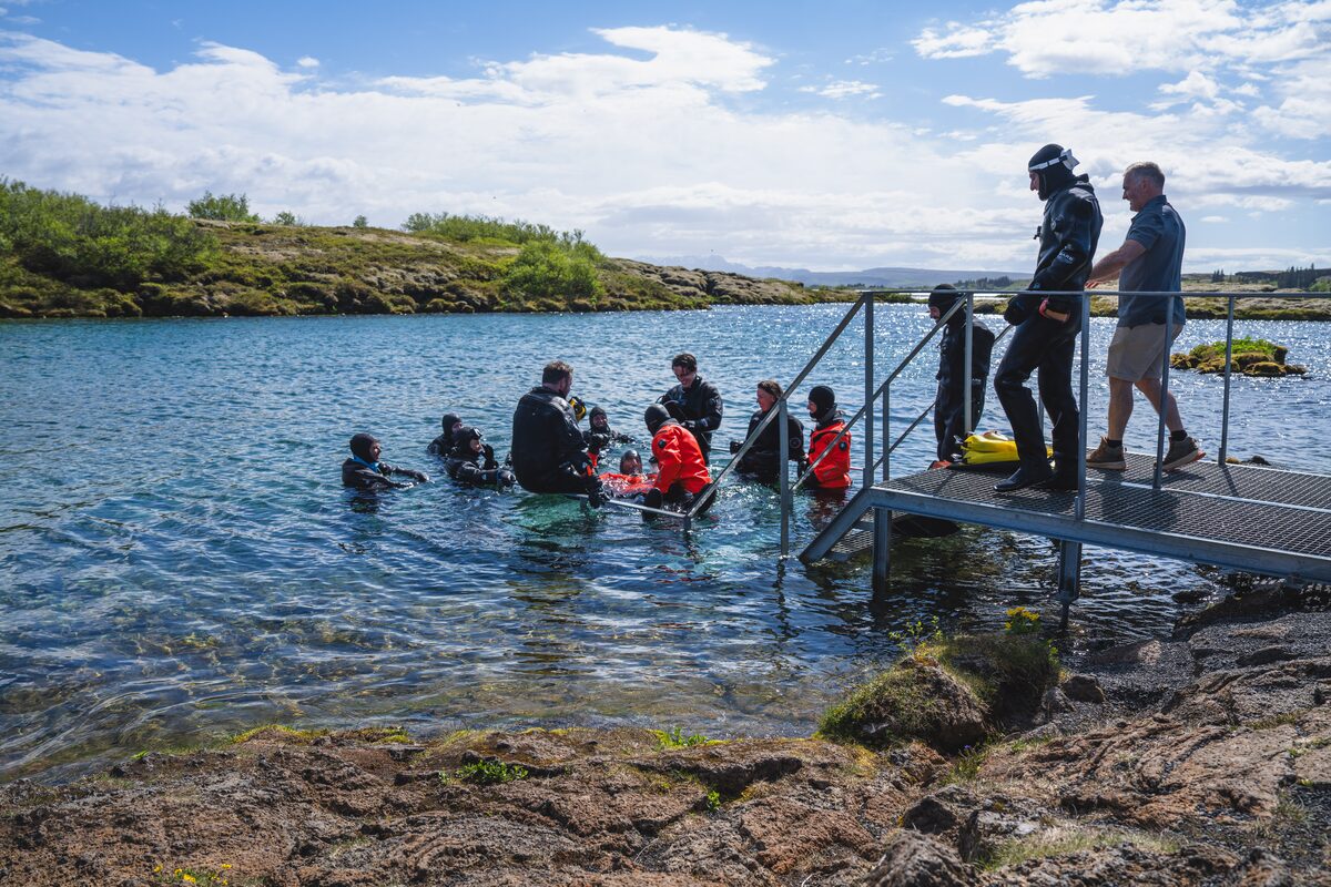 Arctic Adventures guides wearing black and red dry suits practice a cold-water rescue beside the metal entry platform at Silfra, while instructors observe from the shore and platform.