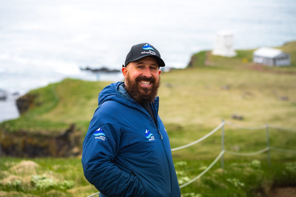 Smiling Arctic Adventures guide in a blue jacket and branded cap standing beside the rugged Westfjords coastline.