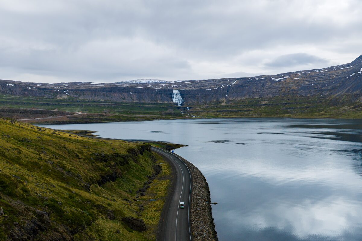 A car on a coastal road beside a calm fjord, with Dynjandi small in the distance below snow-streaked mountains.