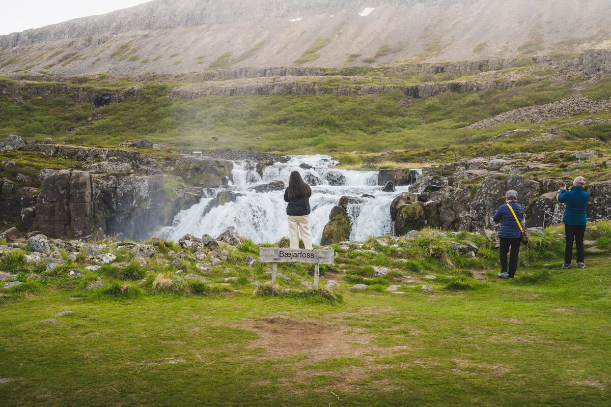 Visitors at the base of a wide low waterfall beside a signpost reading Bæjarfoss, with grassy slopes behind.