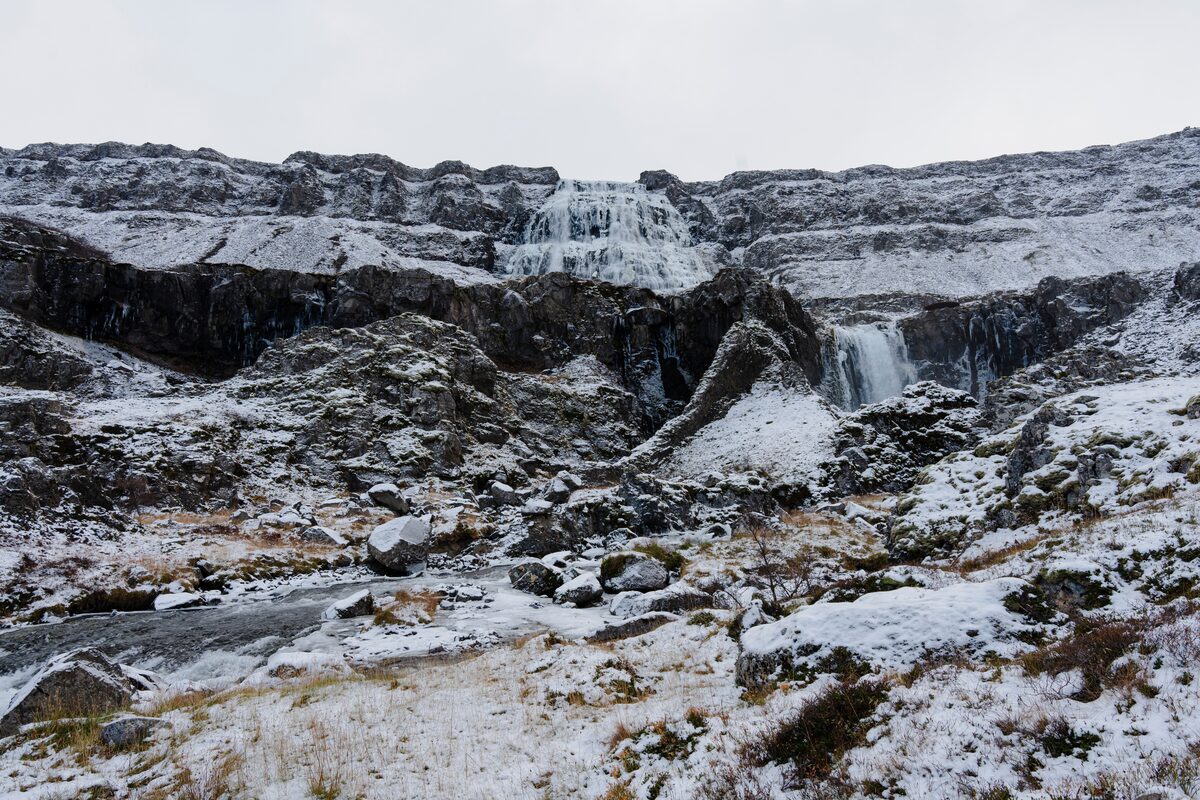 Dynjandi waterfall flowing over snow-dusted rock with thin snow cover on the surrounding slopes and boulders.