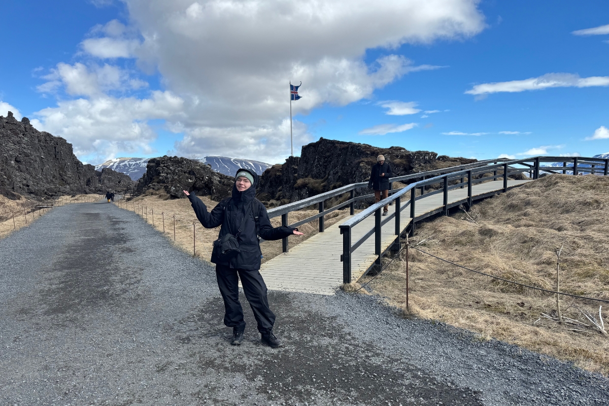 Justyna Zajkovska standing on the gravel path in Þingvellir National Park, with the rift walls and a wooden footbridge beside her.