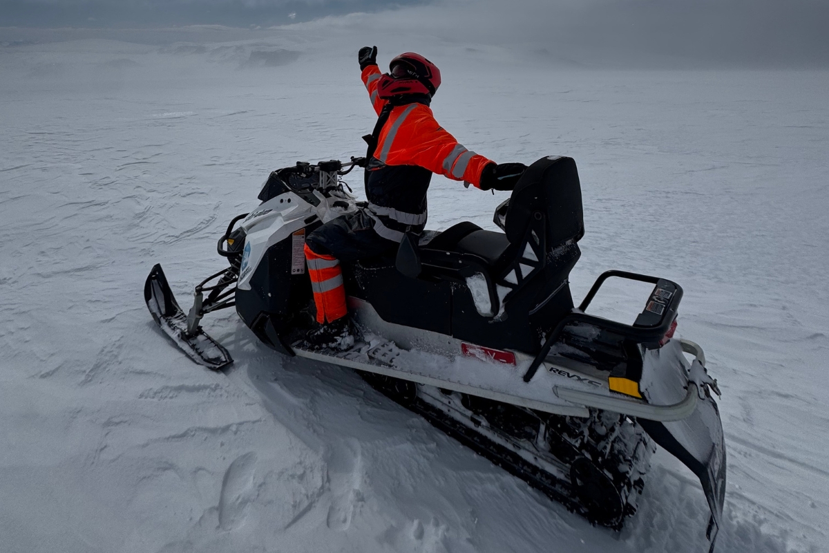 Justyna Zajkovska from Arctic Adventures, snowmobiling on Langjökull glacier in an orange suit and red helmet, one arm raised.